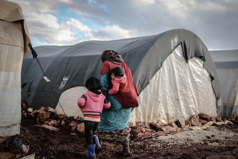 A woman and children walk near tents in a refugee camp in Idlib, showcasing resilience amidst adversity.