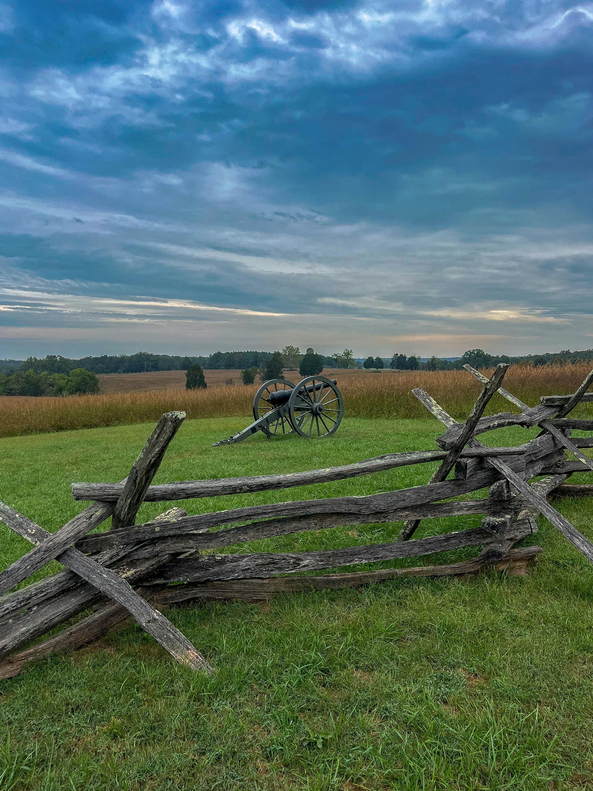 Serene view of a historic Civil War battlefield with cannon and wooden fence under a dramatic sky.