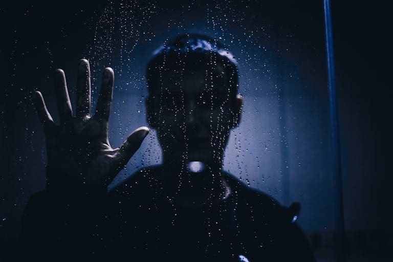 Silhouette of a person with hand against a rain-covered glass, evoking mystery and introspection.